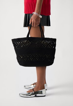 Sac fourre-tout noir en osier avec des motifs découpés, des poignées robustes et un design spacieux. Associé à des chaussures plates argentées et un vêtement texturé.