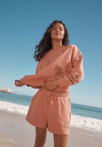 Completo composto da felpa e pantaloncini rosa in cotone con ricami di sole e foglie. Vestibilità comoda con elastico e coulisse in vita.