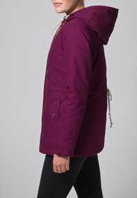 Giacca parka viola aderente con cappuccio, zip frontale, cordoncini e tasche laterali. Realizzata in tessuto testurizzato con una finitura liscia.
