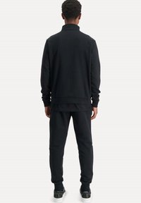 Černý set mikiny a kalhot, vyrobený z měkké bavlněné směsi. Má vysoký límec a manžety, uvolněný střih a žádné viditelné vzory.