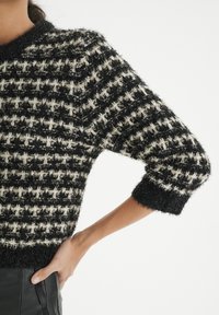 Pull à carreaux noir et blanc avec une texture pelucheuse, manches courtes, et encolure côtelée, associé à un pantalon noir.