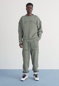 Olivengrønn sweatshirt med ribbede mansjetter og matchende bukser med sidelommer og brodert logo. Bæres med svart og hvit joggesko.