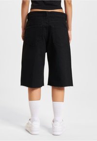 Shorts en denim noir avec ourlets effilochés, deux poches arrière et taille mi-haute. Portés avec des baskets blanches et des chaussettes blanches côtelées.