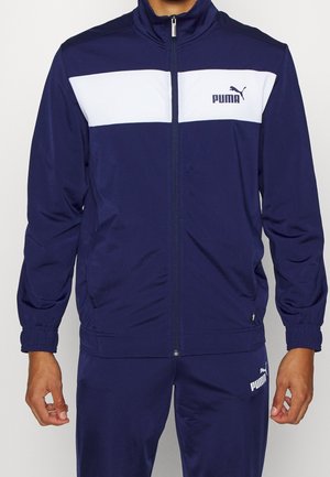 Mann trägt dunkelblauen Puma-Trainingsanzug mit weißem Bruststreifen und Puma-Logo auf Jacke und Hose, steht mit entspannten Armen.