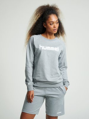 Kvinde med krøllet hår iført en grå hummel sweatshirt og matchende shorts, stående med den ene hånd i lommen og kigger til siden.