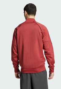 Bordeauxfarbene Sportjacke mit langen Ärmeln, gerippten Bündchen und Saum. Hergestellt aus glattem, dehnbarem Stoff; verfügt über einen subtilen Glanz und einen klaren Schnitt.