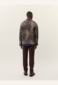 Floral-patterned jacket in shades of purple, beige, and green. Paired with brown pants featuring white side stripes. Textured fabric.