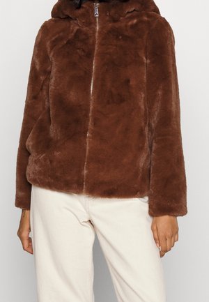 Persona con chaqueta marrón de piel sintética con cremallera, combinada con pantalones blanco roto, vista desde los hombros hasta las caderas sobre un fondo liso.