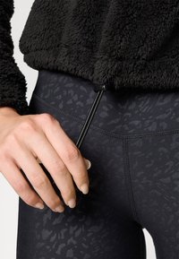 Pull en polaire noir avec une finition texturée, resserré à l'ourlet par un cordon de serrage noir. Porté par-dessus un legging noir à motifs.