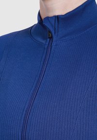 Close-up van een persoon die een blauw ribbelig jasje met rits en een hoge kraag draagt, die tot aan de nek is dichtgeritst.