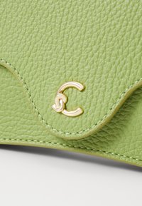 Borsa in pelle verde con texture, bordi scallopati e un logo dorato. Presenta cuciture visibili e una finitura liscia.