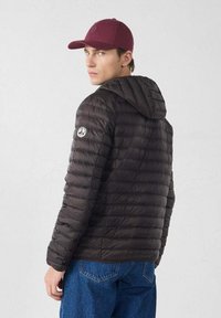 Uomo che indossa una giacca nera trapuntata con cappuccio e un berretto color bordeaux, che guarda oltre la spalla su uno sfondo chiaro e uniforme.