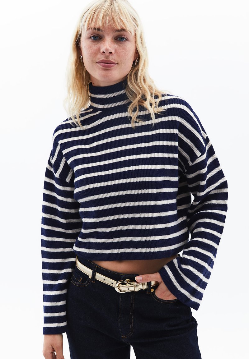 Randig stickad tröja med hög krage, med marinblå och krämfärgade horisontella ränder, kort design och ribbad textur. Kompletterar mörka jeans.