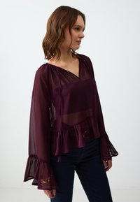 Blusa trasparente bordeaux con scollatura a V, orlo arricciato e maniche a campana, con lacci sul colletto e una texture leggera.
