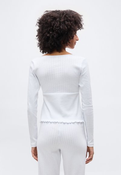 Γυναίκα με σγουρά μαλλιά που κοιτάζει πλάγια, φοράει λευκό μακρυμάνικο τοπ και παντελόνι με διακριτικά πουά μοτίβα.