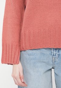 Rosa strikket genser med ribbestrikket kant, med løse ermer og teksturert overflate. Kombinert med lyseblå denimjeans.