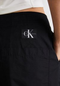Crne hlače s vidljivom oznakom brenda Calvin Klein Jeans. Tkanina izgleda glatko s strukturiranim dizajnom i bočnim džepovima.
