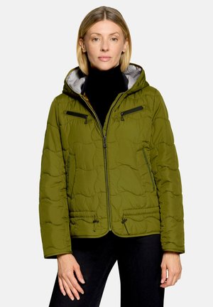 Femme aux cheveux blonds courts portant une veste matelassée verte à capuche par-dessus un col roulé noir et un pantalon noir sur un fond uni.