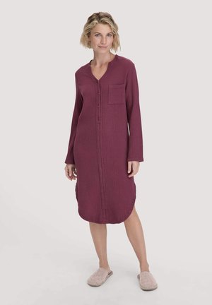 Donna in piedi che indossa un vestito da notte bordeaux con maniche lunghe e bottoni, dotato di una tasca frontale, e pantofole chiare su uno sfondo semplice.
