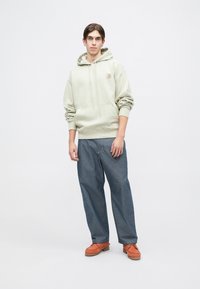 Sudadera de algodón verde claro con un bolsillo frontal, combinada con pantalones de mezclilla oscuros de corte holgado y zapatos naranjas. De pie contra un fondo liso.