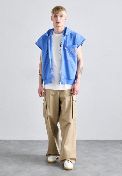 Himmelblaues ärmelloses Hoodie mit Frontreißverschluss, seitlichen Taschen und Logodetail. Kombiniert mit beigen Cargo-Hosen und weißen Sneakers.