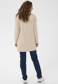 Ženska z valovitimi rjavimi lasmi, oblečena v bež tuniko z dolgimi rokavi, temno modre kavbojke in bele superge, ki stoji proti enobarvni ozadju.