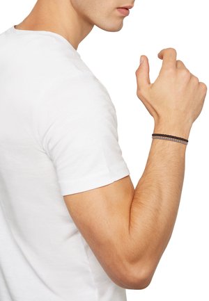 Sort flettet læderarmbånd med sølvaccent, båret på en bar håndled. Personen har en simpel hvid t-shirt på.