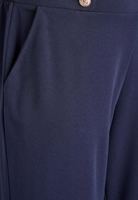 Detail tmavě modrých kalhot s boční kapsou a zlatým knoflíkem na pasu s motivem kotvy.