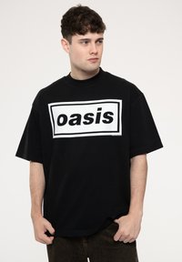 Black oversized t-shirt made of cotton, featuring a bold white rectangular graphic with "oasis" in black lettering. Short sleeves, round neck.