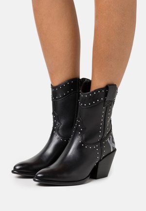 Jambes portant des bottines cowboy en cuir noir avec des clous argentés et des talons carrés sur fond blanc.