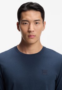 Marineblaues Langarmshirt aus Baumwolle mit rundem Ausschnitt und einem quadratischen Logoetikett auf der linken Brust. Glatte Textur.