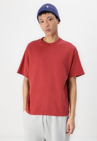 Tricou roșu din bumbac, cu mâneci scurte și guler rotund, stilizat cu o pereche de pantaloni de trening gri. O căciulă albastră cu model fălcat completează ținuta.