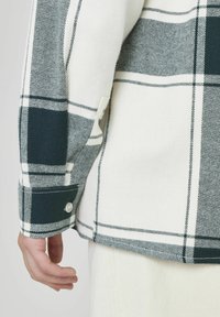 Chemise en flanelle à carreaux blancs et verts foncés, avec des poignets à boutons et des textures contrastées, des coutures visibles et un détail de poche.