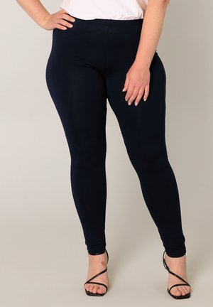 Vrouw draagt donkerblauwe leggings en zwarte sandalen met bandjes en hoge hakken, staand tegen een effen lichte achtergrond.