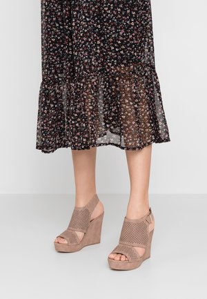 Nogi osoby ubrane w czarną, kwiatową spódnicę midi oraz beżowe, ażurowe sandały na koturnie z zapięciem na kostce, stojące na jasnej podłodze.