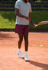 Tennis Spieler in einem hellgrauen Shirt und maronenfarbenen Shorts, bereit mit einem Schläger auf dem Sandplatz, mit einem sich in der Nähe befindlichen gelben Tennisball in Bewegung.