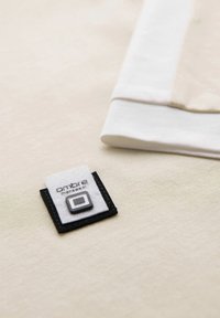 Světlý béžový materiál s hladkou texturou; bílá štítek s černými a šedými akcenty, na kterém je výrazně napsáno "ombre menswear".