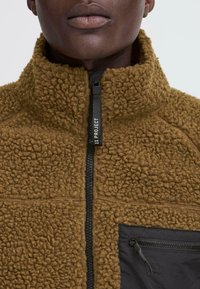 Close-up of a person wearing a brown textured fleece jacket with a black zipper and chest pocket, featuring a pull tab labeled "11 PROJECT".