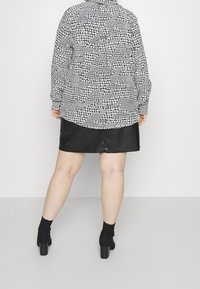 Chemise à boutons noire et blanche à motifs, à manches longues, associée à une mini-jupe en cuir noir. Des chaussettes noires et des escarpins épais complètent la tenue.
