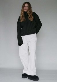 Czarny prążkowany sweter z szerokimi rękawami, zestawiony z białymi spodniami z wysokim stanem. Czarne buty na platformie dopełniają stylizację. Neutralne tło.
