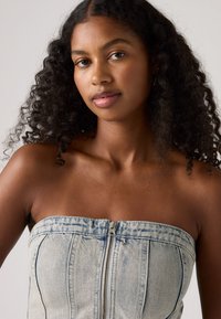 Top in denim senza spalline di colore azzurro chiaro con zip frontale. Presenta cuciture verticali e una finitura liscia e strutturata. Nessun ulteriore dettaglio decorativo.