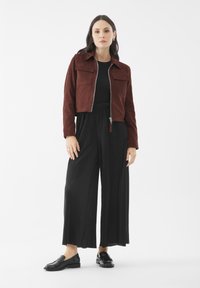 Hnědá semišová zkrácená bunda se dvěma náprsními kapsami, spárovaná s černými širokými kalhotami s plisováním a černými mokasíny. Minimalistický design, hladká textura.