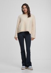 Hellbeigefarbener, fluffiger Pullover mit lockerer Passform und weiten Bündchen, kombiniert mit dunkelblauen Schlaghosen und grauen Sneakers. Glatte Textur und einfaches Design.