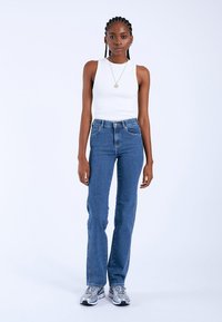 Top bianco aderente e jeans blu a vita alta, con taglio dritto. Indossato con scarpe da ginnastica argentate, in piedi contro uno sfondo neutro.
