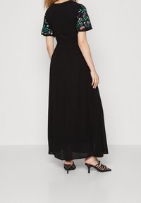 Schwarzes Maxikleid mit kurzen, bestickten Blumenärmeln; leichtes Material; fließende Silhouette; kombiniert mit schwarzen, geschlossenen Sandalen mit Absatz.