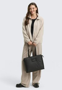 Sac fourre-tout noir rembourré avec des poignées doubles et une surface texturée. Présente une étiquette à l'avant. Porté avec une veste en polaire légère et un pantalon large.
