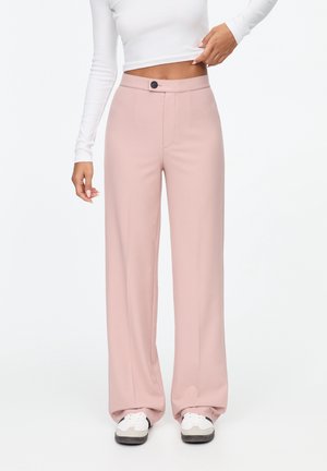 Kvinde iført høj talje, vidde rosa bukser, hvid langærmet cropped top og hvide sneakers, stående mod en hvid baggrund.