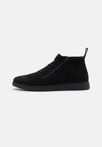 Las botas de tobillo de ante negro cuentan con una punta redondeada, cordones frontales y una cremallera lateral. La suela es plana con un acabado texturizado para un mejor agarre.