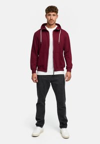Burgundy zip-up hoodie with a drawstring hood, front pockets, and elastic cuffs, worn over a white t-shirt and paired with dark jeans.