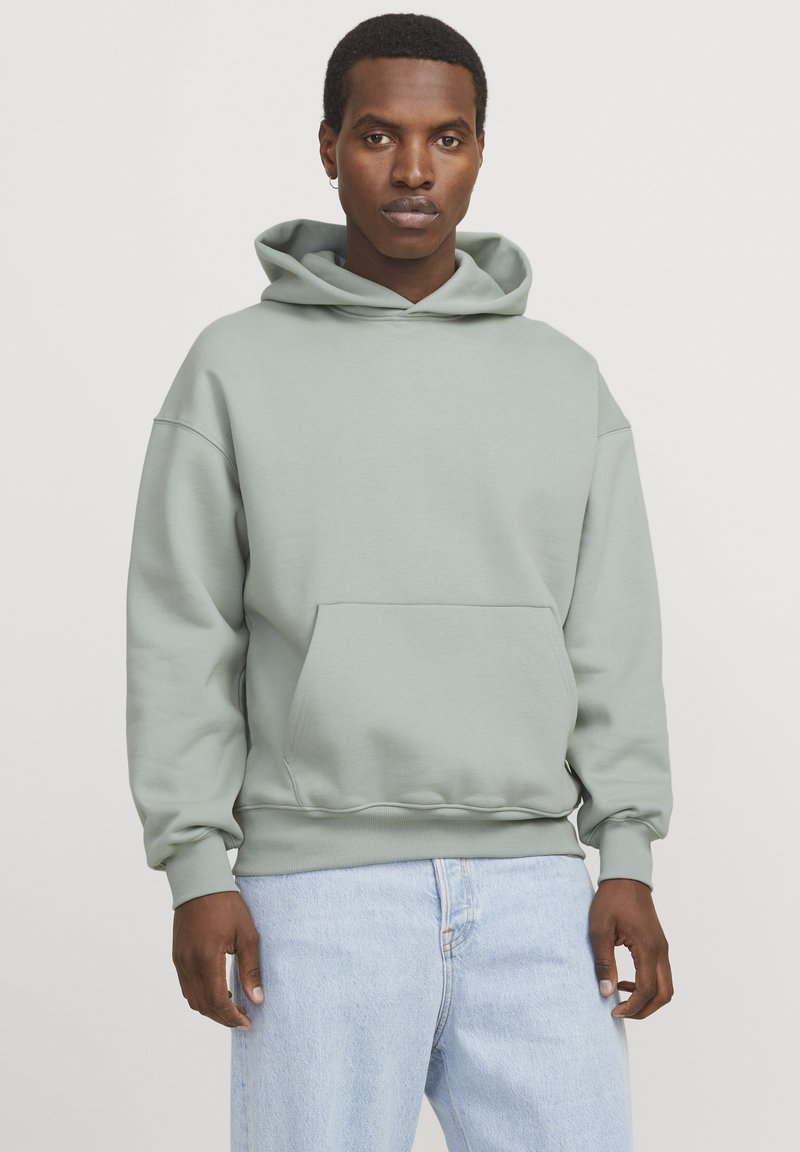 Felpa con cappuccio verde chiaro, realizzata in tessuto morbido. Presenta una tasca frontale, polsini a costine e orlo. Indossata con jeans azzurri.